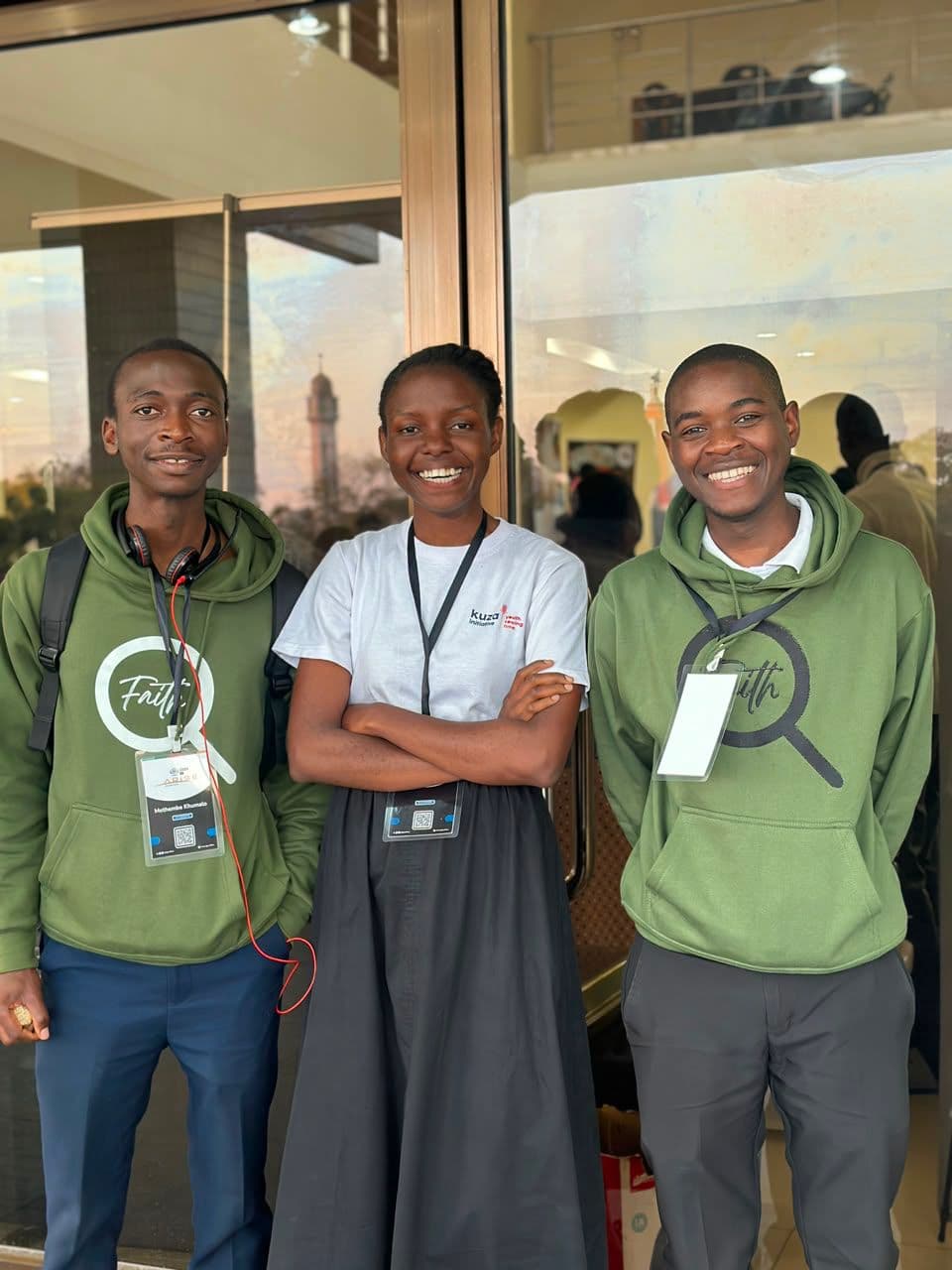 Three smiling conference attendees outside the venue, two wearing green Faith hoodies